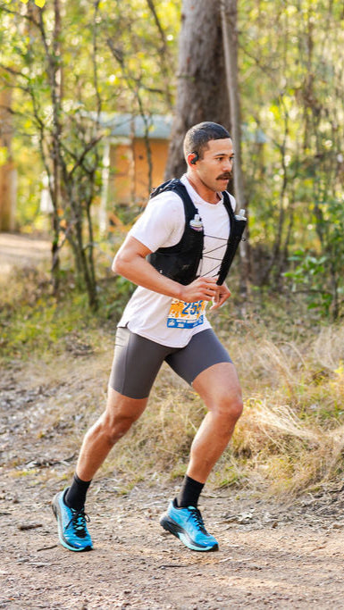 Man running on a trail in a forested area with a visible brand logo.